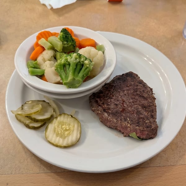 A plate with a grilled steak, a side of steamed vegetables, and sliced pickles.